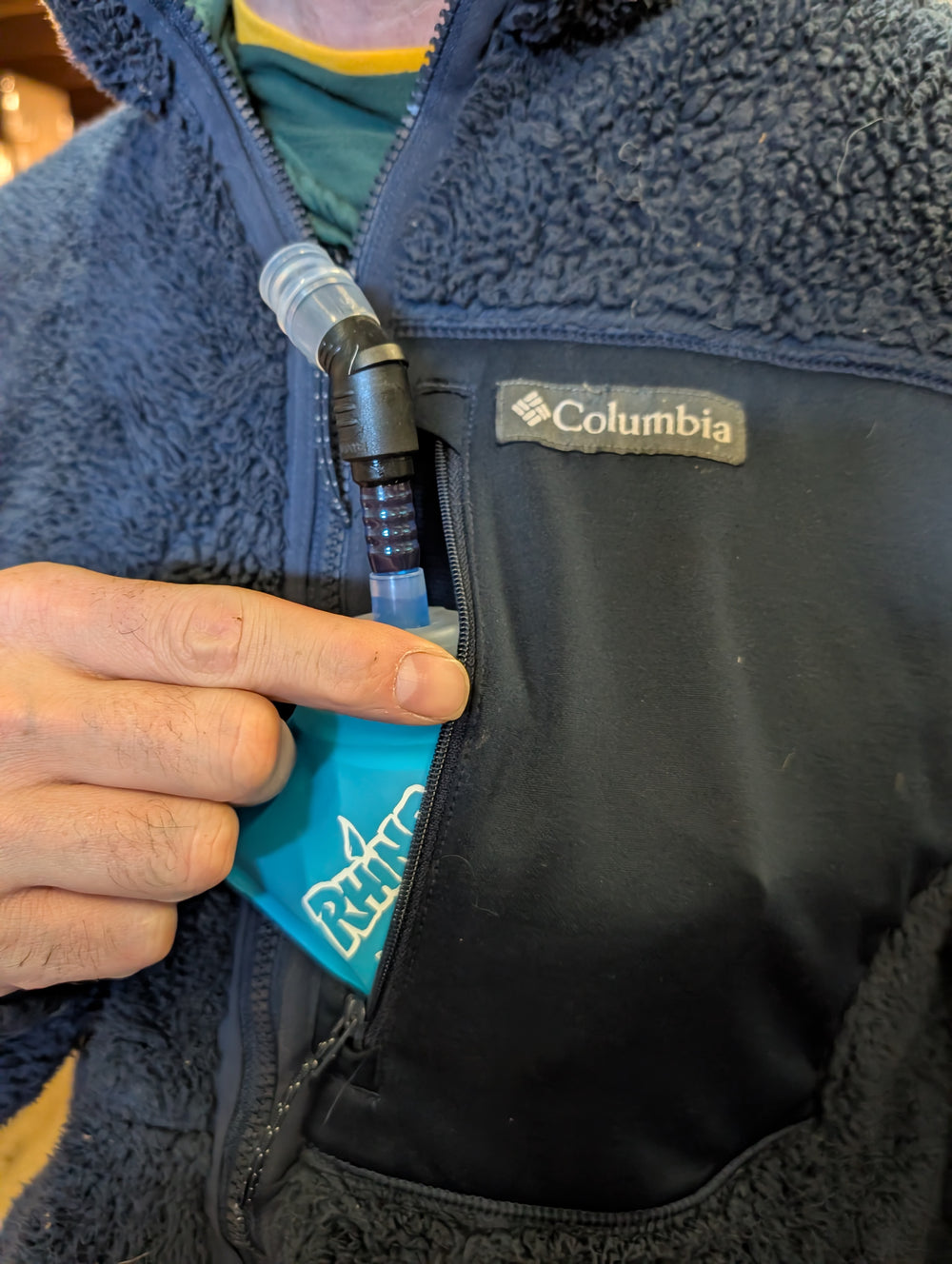 A close-up of a person's hand inserting a blue and black hydration tube into a zippered pocket of a dark blue fleece Columbia jacket.
