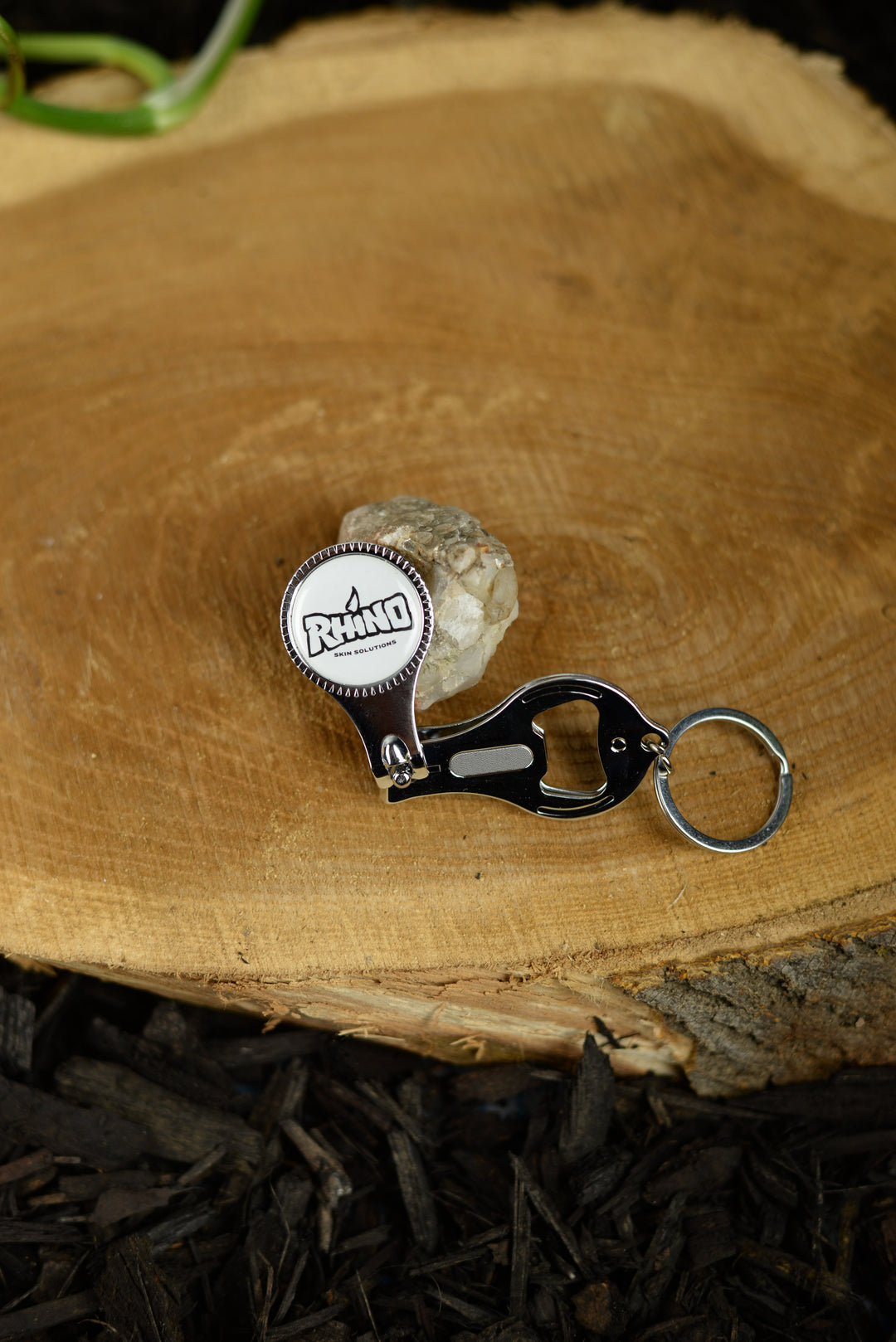 A Rhino-branded nail clipper with an integrated bottle opener fully opened, revealing the bottle opener feature, resting on a log. The metal opener stands out against the rough, natural texture of the wood, showcasing its practical design in an outdoor setting. The Rhino logo is visible on the clipper, emphasizing the brand's focus on rugged, multifunctional tools ideal for outdoor enthusiasts. The contrast between the polished metal and the raw log surface highlights the tool's durability and versatility.
