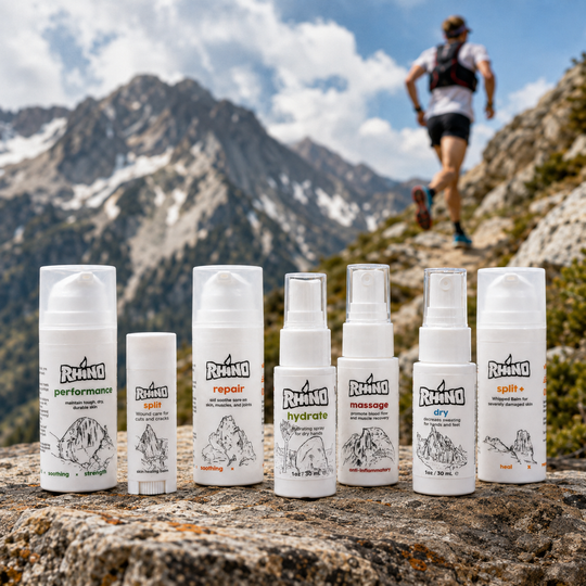 Seven white outdoor care products from Rhino are lined up on a rocky mountain path, with a blurred hiker in the background against snow-capped peaks and blue sky.