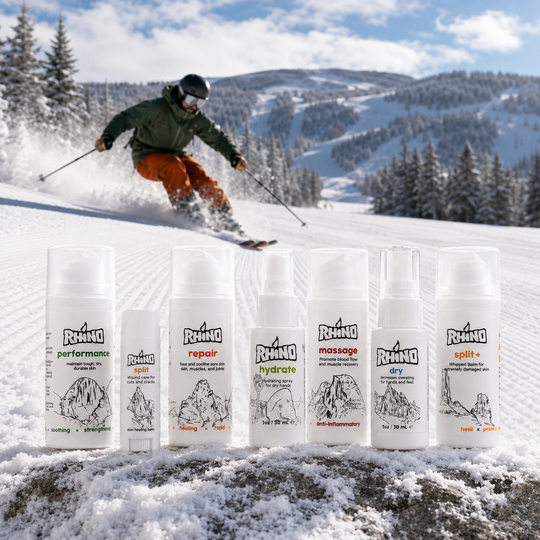 Six white Rhino ski and snowboard care bottles and sprays are lined up in the snow, with a skier carving through powder in the background on a mountain slope.