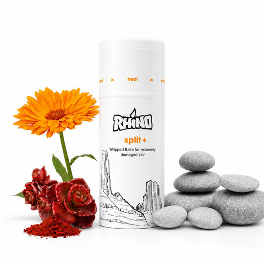 A white cylindrical bottle of RHINO split+ whipped balm for damaged skin, styled on a white background with an orange marigold flower, red rose, red powder, and gray decorative stones.