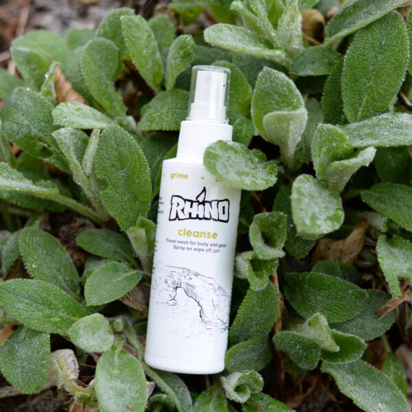 A Cleanse Spray bottle nestled in a bed of fresh herbs. The natural setting emphasizes the product's infusion of herbal ingredients like tea tree oil, peppermint, and lavender. The green herbs surrounding the bottle highlight its connection to nature and the use of organic, skin-soothing elements in its formulation. The arrangement showcases the spray as a refreshing, natural solution for cleaning and revitalizing both skin and gear after outdoor activities.