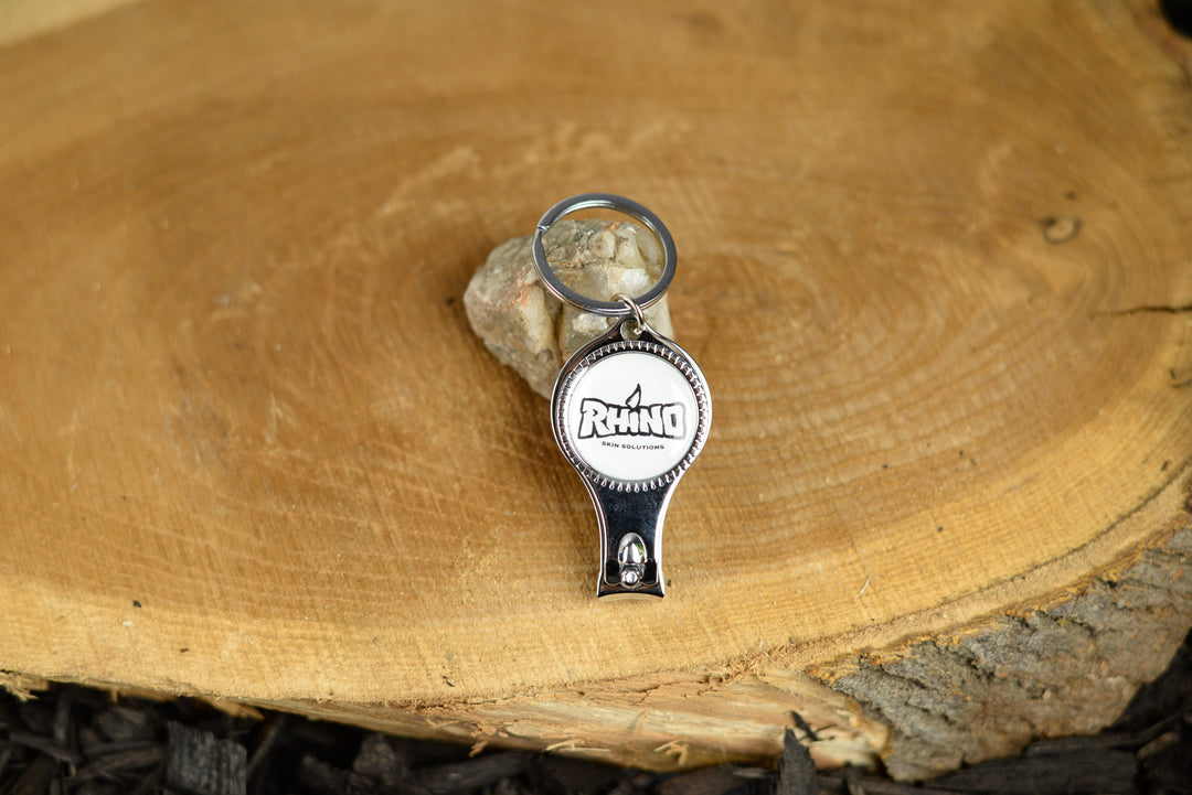 A Rhino-branded nail clipper with an integrated bottle opener placed on a tree stump. The clipper, featuring the Rhino logo, rests securely on the rough, natural surface of the stump, contrasting its polished metal finish with the rugged texture of the wood. The setting highlights the tool's durability and outdoor utility, making it an ideal accessory for adventurers and nature enthusiasts. The natural backdrop emphasizes the blend of practicality and style in this multifunctional tool.